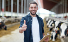 agriculture industry, farming, people and animal husbandry concept - happy smiling young man or farmer with clipboard and cows in cowshed on dairy farm showing thumbs up hand sign
